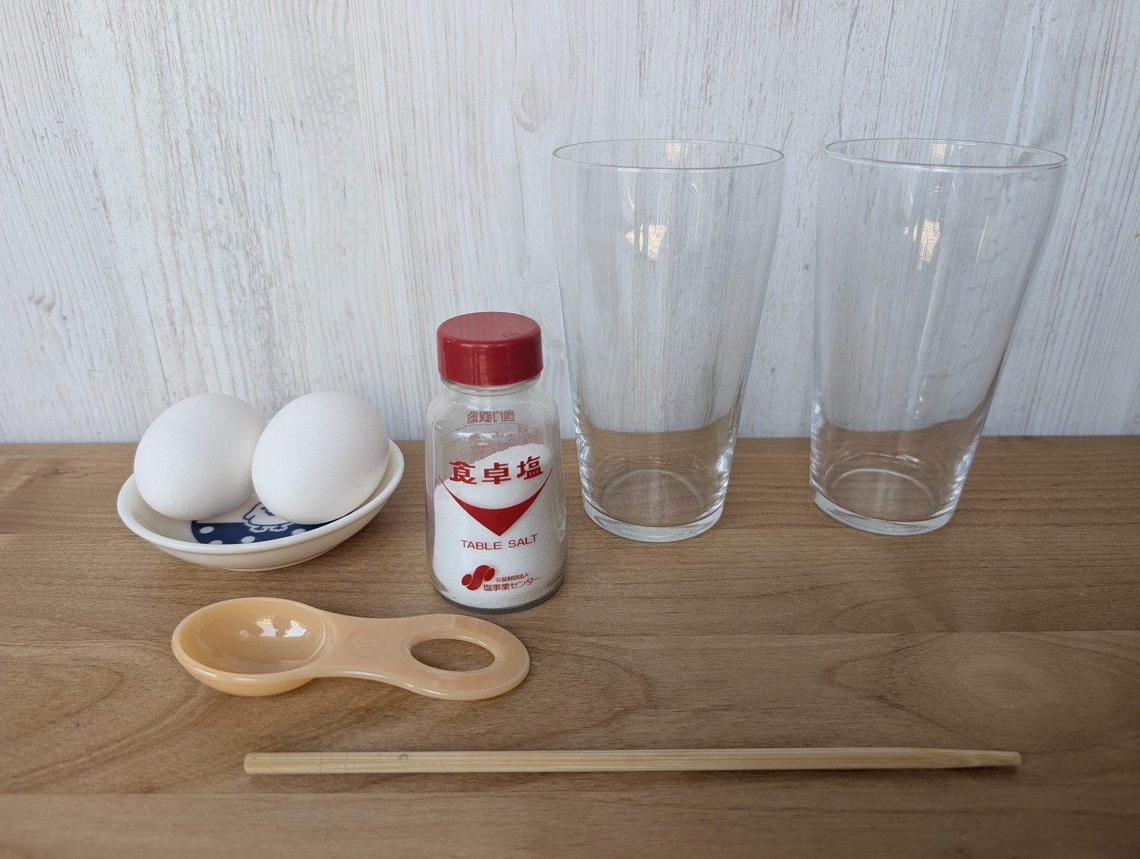 卵2個・食卓塩・計量スプーン・菜箸・透明なコップ2個を木のテーブルに並べた写真